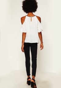 Vit cold-shoulder topp med en nyckelhålsdetalj på ryggen, lättviktsmaterial och vågig fåll; matchad med svarta smala jeans och högklackade skor.