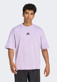 Fialové tričko s krátkým rukávem vyrobené z měkké tkaniny, ve středu s černým logem Adidas. Jednoduchý, uvolněný střih.