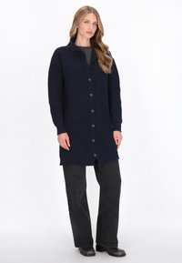 Granatowy sweter robiony na drutach z teksturowanym wzorem, z wysokim kołnierzem, guzikami z przodu i długimi rękawami, połączony z czarnymi spodniami dzwonami.