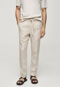Beige linnen broek met een relaxte pasvorm en een koord in de taille; gecombineerd met een lichtgrijze top met korte mouwen en zwarte sandalen.