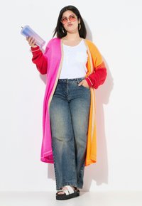 Cárdigan largo colorido con secciones en rosa, naranja y rojo. Top blanco con un bolsillo en el pecho. Jeans de mezclilla azules de cintura alta y sandalias negras gruesas.