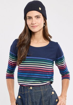 Donkerblauwe langemouwen shirt met kleurrijke horizontale strepen in groen, blauw, roze en crèmekleur; gedragen met een donkere beanie en denim broek.