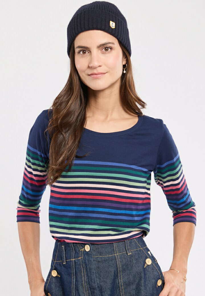 Camicia a maniche lunghe blu navy con strisce orizzontali colorate in verde, blu, rosa e crema; indossata con un berretto scuro e pantaloni in denim.