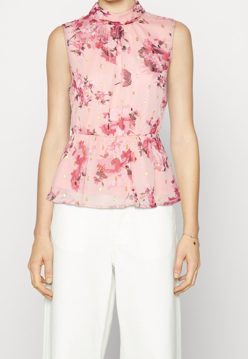 Blouse sans manches rose à fleurs avec des accents dorés, col montant, taille peplum, portée avec un pantalon blanc et un bracelet délicat au poignet droit.