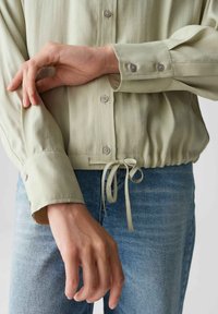 Lichtgroene, lange mouwen shirt met knoopmanchetten en drawstring zoom. Glad textuur en subtiele glans; gecombineerd met blauwe denim jeans.