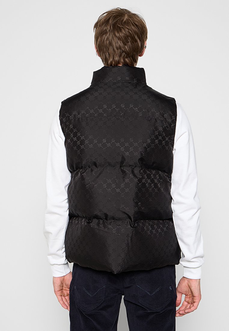 Zwarte gewatteerde vest met een subtiele reliëfpatroon, voorzien van een hoge kraag en een gladde textuur. Ontwerp met open rug, gedragen over een wit shirt.