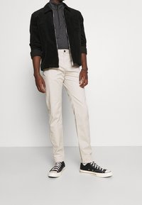 Zwarte korduroy zipjas, grijze getailleerde coltrui, crèmekleurige rechte broek, zwarte sneakers met witte zolen en subtiele kantdetails.