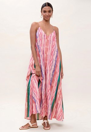 Mujer vestida con un maxi vestido a rayas de colores rosa, púrpura y verde, con tirantes finos y sandalias beige, de pie frente a un fondo liso.