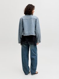 Άτομο με σγουρά μαλλιά, γυρισμένο πλάτη, φοράει ανοιχτό μπλε τζιν μπουφάν πάνω από μαύρη μπλούζα και φαρδιά μπλε τζιν, με φόντο απλό.