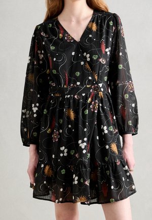 Frau trägt schwarzes, langärmliges Wickelkleid mit bunten abstrakten Blumen- und Handmustern, geraffter Taille und Rüschensaum.