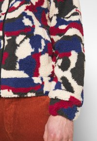 Fleecejacka med ett mångfärgat abstrakt mönster i kräm, blått, rött och svart; texturerad, med dragkedja och elastiska ärmslut.