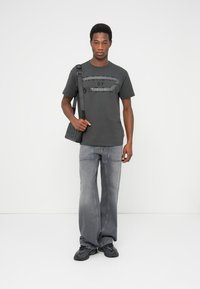 Maglietta grigio scuro con dettaglio logo, abbinata a jeans grigi a gamba larga e sneakers nere. La persona porta una borsa a tracolla nera.