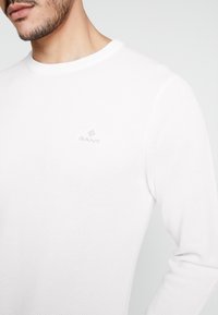 Weißes langärmeliges Strickshirt mit rundem Halsausschnitt, das aus dezent strukturiertem Material besteht und ein kleines, graues GANT-Logo auf der Brust trägt.
