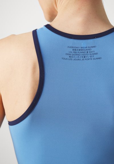 Blå ärmlös topp med marinblå fåll, med tryckt text på flera språk på baksidan. Slät textur och figurnära design.