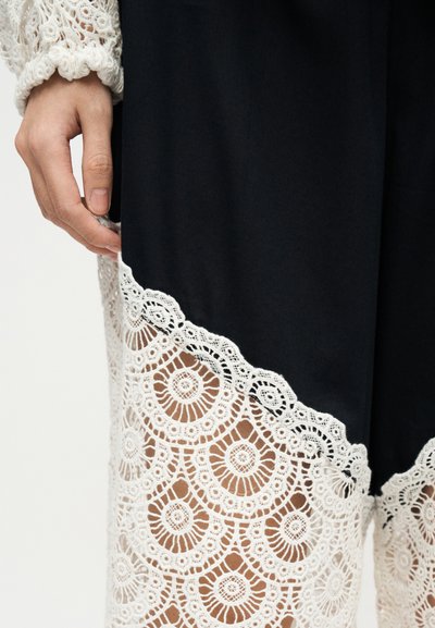 Main posée à côté d'un tissu noir avec un bord en dentelle blanche complexe et une manche en dentelle détaillée visible sur un fond clair.