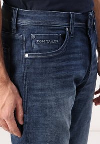 Dark blue denim jeans with a slim fit; features a front pocket with a logo and silver hardware; visible stitching and texture details.