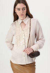 Blusa rosa chiaro con un leggero lucido, caratterizzata da un intricato ricamo dorato sulla parte anteriore, chiusura con bottoni e maniche lunghe con polsini decorati.