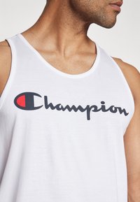 Top sin mangas de algodón blanco con un destacado logo en negro y rojo de "Champion". La tela parece suave con un clásico escote redondo.