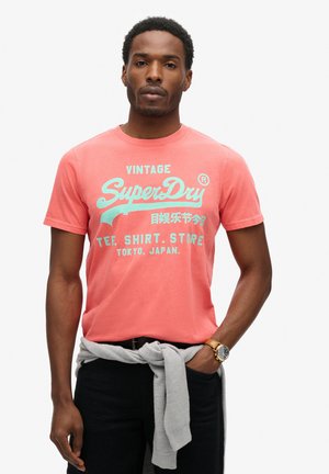 Coral cotton t-shirt featuring turquoise "SuperDry" logo and text. Short sleeves, round neckline, worn with a grey sweater tied at the waist.