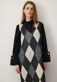 Egyenes vörös hajú nő fekete hosszú ujjú inget visel ujjatlan, fekete, fehér és szürke rombusz mintás ruhája alatt.