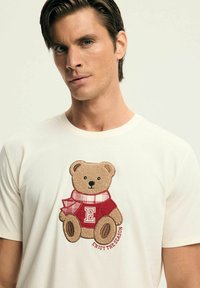 Kremowy bawełniany t-shirt z haftowanym misiem w czerwonym swetrze z kratkowanymi akcentami, z napisem "Ciesz się sezonem."