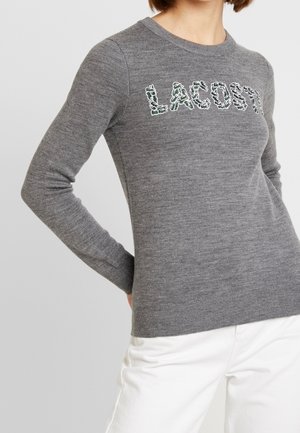 Persoon draagt een grijze Lacoste trui met lange mouwen met een wit- en groen patroonlogo en witte broek tegen een effen achtergrond.