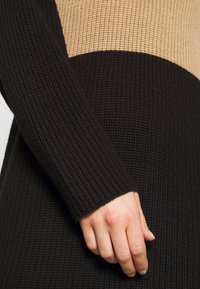 Gros plan sur une main reposant sur le côté, portant une robe tricotée à manches longues avec des blocs de couleur beige et noir.