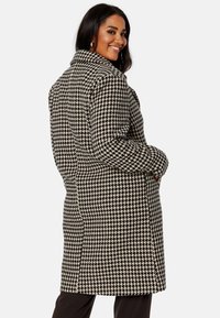 Cappotto a motivo pied-de-poule in bianco e nero con colletto a disfida, maniche lunghe e tasche laterali, che presenta una silhouette sartoriale.