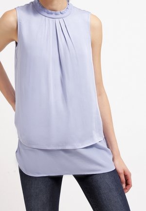 Ærmeløs lyse lilla bluse med opsamlede læg under en flæsekrave, lagdelt kant, båret med mørkeblå jeans.