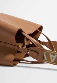 Tan leather handbag with a soft texture, featuring gold hardware, and a patterned shoulder strap. Open top design shows interior compartments.