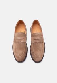 Ζευγάρι από ανοιχτό καφέ μοκασίνια από σουέντ με στρογγυλεμένη μύτη και λεπτομέρεια με λουράκι, τοποθετημένα δίπλα-δίπλα σε λευκό φόντο.