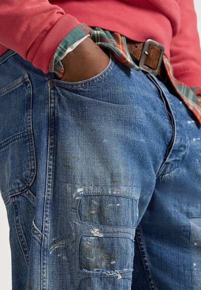 Blå denim cargobukser med synlige malingsflekker, med flere lommer og en avslappet passform. Brukt med en rød genser over en rutete skjorte.