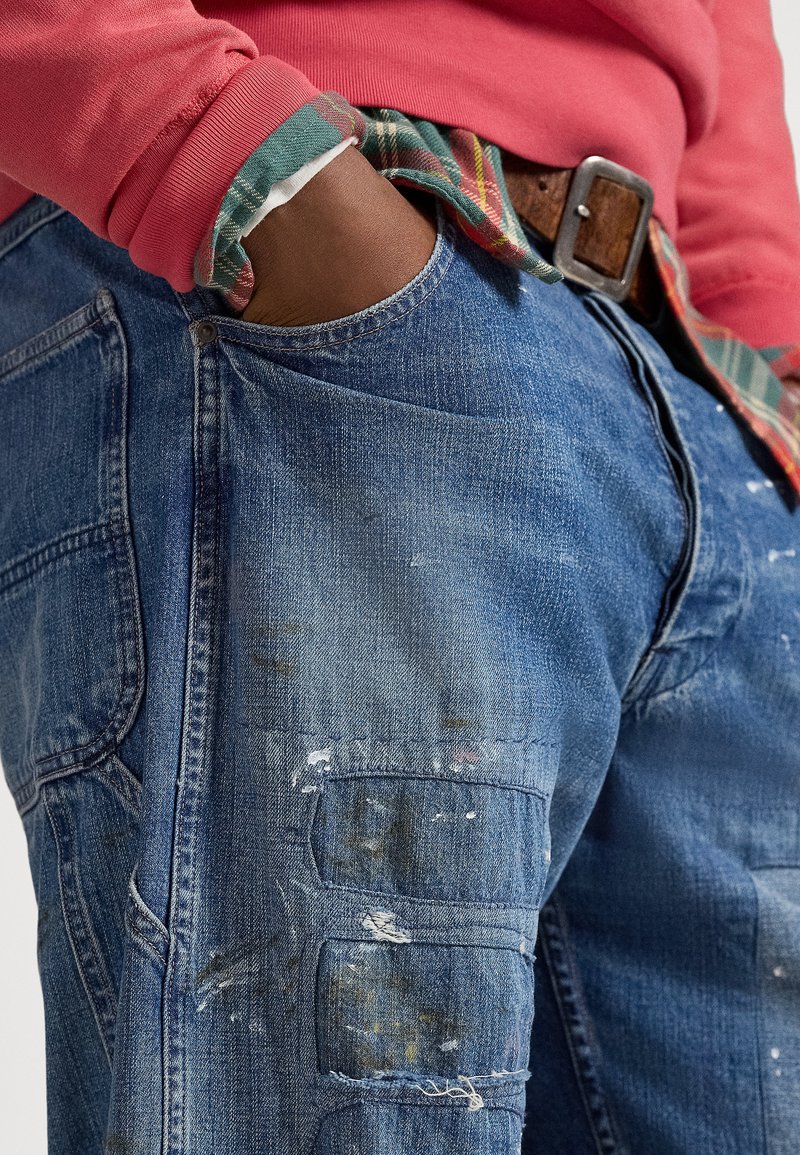 Pantaloni cargo in denim blu con macchie di vernice visibili, dotati di molteplici tasche e vestibilità comoda. Indossati con una felpa rossa sopra una camicia a quadri.