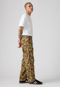 Pantalones cargo con camuflaje en tonos marrones y verdes, con múltiples bolsillos, combinados con una camiseta blanca y zapatillas negras con rayas blancas.