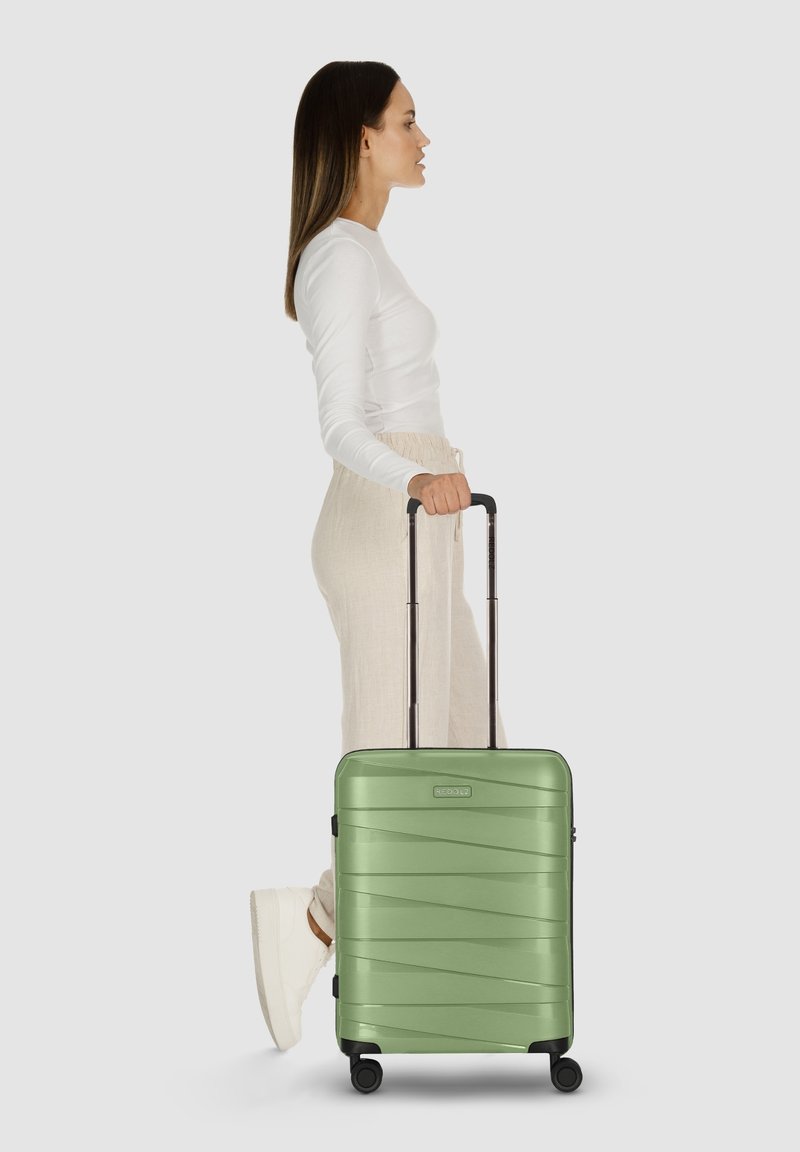 Donna con top bianco e pantaloni beige che cammina con una valigia a rotelle verde su uno sfondo semplice.
