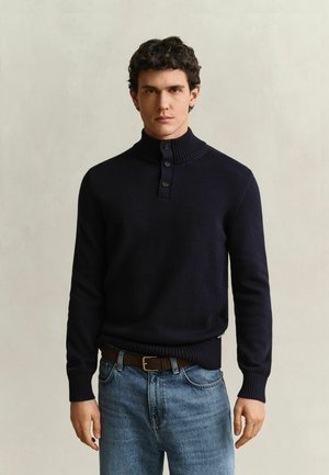 Jeune homme aux cheveux bouclés et foncés, portant un pull bleu marine boutonné et un jean bleu, debout devant un fond clair uni.