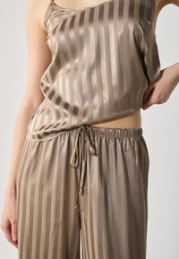 Silkkisekoite loungewear-setti vaaleanruskeana, jossa on pystysuorat raidat. Asusteeseen kuuluu väljä toppi ja nyörihaalarit, joissa on elastinen vyötärö.