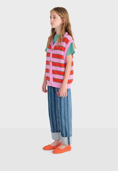 Niña joven de perfil que lleva un chaleco de punto a rayas rosas y rojas, camiseta verde, vaqueros remangados azules y zapatos naranjas sin cordones.