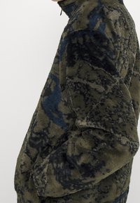 Fleecejacke in Dunkelgrün mit blauen und schwarzen abstrakten Mustern. Hoher Kragen, Reißverschluss und weich strukturierter Stoff.