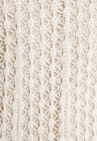 Tissu tricoté couleur crème avec un motif en losange en dentelle superposé sur un matériau beige, créant un aspect texturé et semi-transparent.