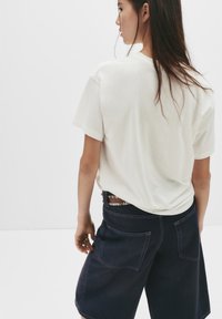 Kvinde med langt mørkt hår iført en hvid t-shirt og mørke denim shorts med en nitterbælte, set bagfra mod en hvid baggrund.