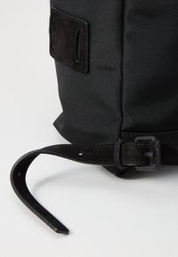 Sort stofrygsæk med en tekstureret overflade, der har en suedestrop og et rektangulært mærke. Spænde lukning med et robust design.