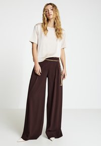 Top beige chiaro a maniche corte abbinato a pantaloni larghi marrone scuro, con cintura a catena dorata e scarpe color crema con punta a punta.