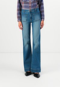 Kvinne som har på seg blå høyt liv flared jeans med store frontlommer, en lilla rutete skjorte og svarte spisse sko.