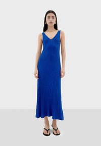 Robe maxi bleue à côtes avec un décolleté en V, corsage ajusté et ourlet fluide. Le mannequin porte des tongs noires et un collier de perles colorées.