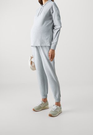 Gravid person iført lysegrå hættetrøje og sweatpants med beige sneakers, der holder en lille cremefarvet håndtaske ved siden af sig.