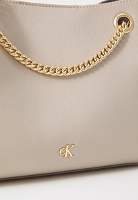 Šviesiai ruda odinė rankinė su tekstūruota paviršiumi, su auksine grandinės dirželiu ir auksiniu "CK" logotipu priekyje.
