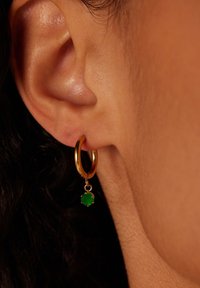 Gouden creolen met een kleine groene stenen hanger, hangend aan de creolen, tegen een close-up van de oorlel en huid.