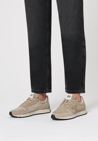 Sneakers in suede beige chiaro con suola bianca e sottili accenti neri a contrasto. Presentano un design con lacci e un logo sulla linguetta.
