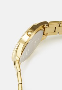 Goldfarbene Armbanduhr mit band aus poliertem Metall, runder Gehäuseform und runder Krone. Besitzt eine glatte, reflektierende Oberfläche. Die Rückseite ist silberfarben.